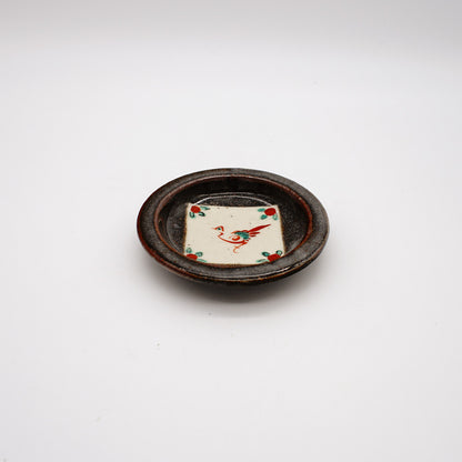 Small round ceramic dish with dark rustic exterior and white center featuring hand-painted red crane design, showcasing traditional Japanese pottery techniques