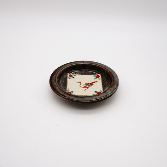 Small round ceramic dish with dark rustic exterior and white center featuring hand-painted red crane design, showcasing traditional Japanese pottery techniques